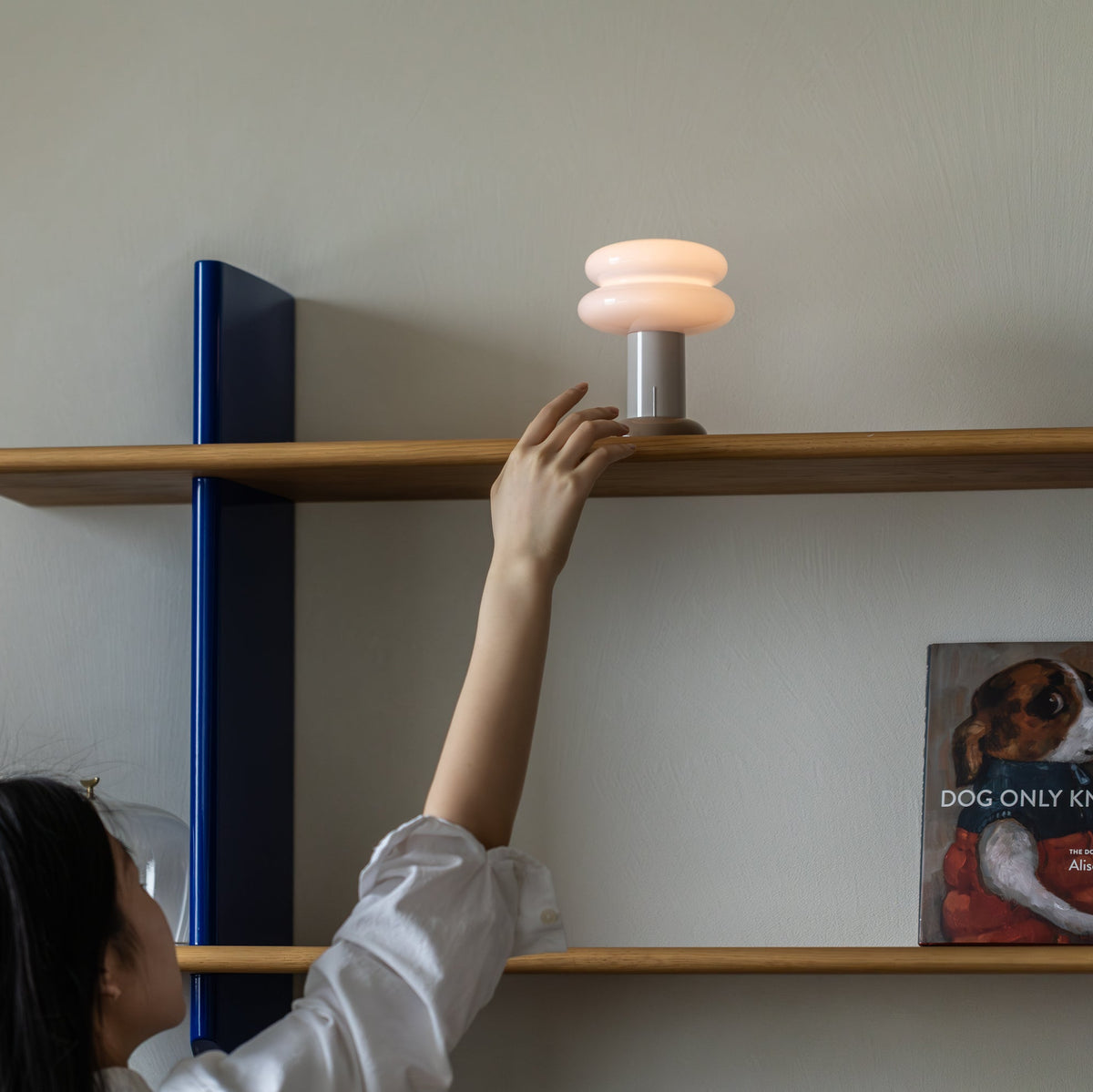 A person reaches up to turn on a modern stacked-shelf table lamp with a glowing warm white shade, mounted on a wooden shelf with blue side brackets.
