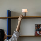 A person reaches up to turn on a modern stacked-shelf table lamp with a glowing warm white shade, mounted on a wooden shelf with blue side brackets.