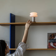 A person reaches up to turn on a modern stacked-shelf table lamp with a glowing warm white shade, mounted on a wooden shelf with blue side brackets.
