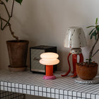 A modern mushroom-shaped table lamp with a glowing white rounded shade and a pink base sits on a tiled shelf next to a black Marshall speaker, a red octopus lamp, and potted plants.