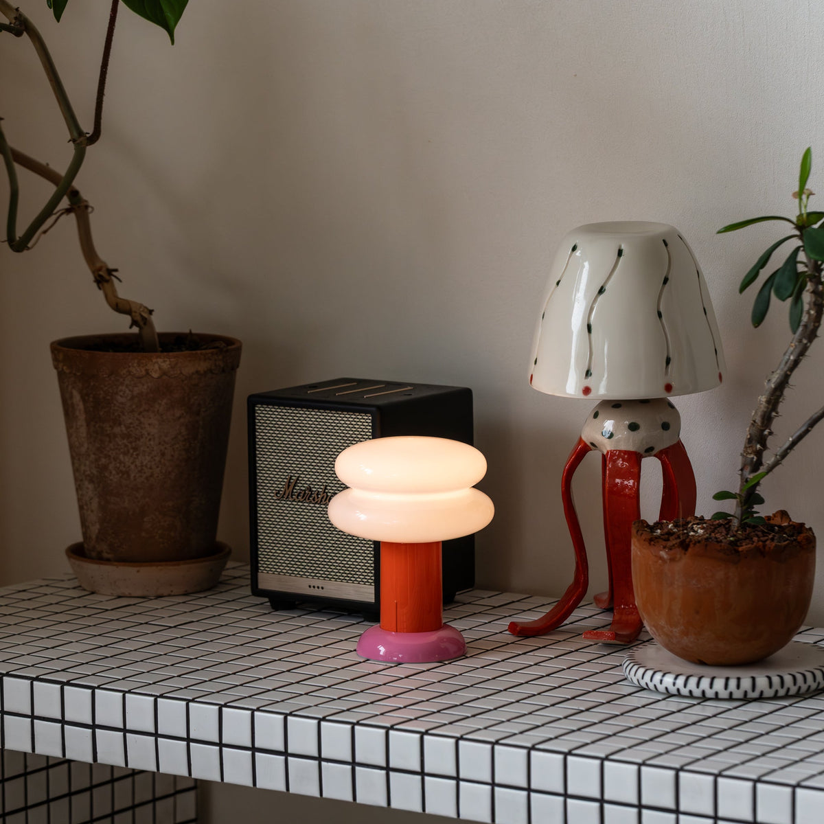 A modern mushroom-shaped table lamp with a glowing white rounded shade and a pink base sits on a tiled shelf next to a black Marshall speaker, a red octopus lamp, and potted plants.