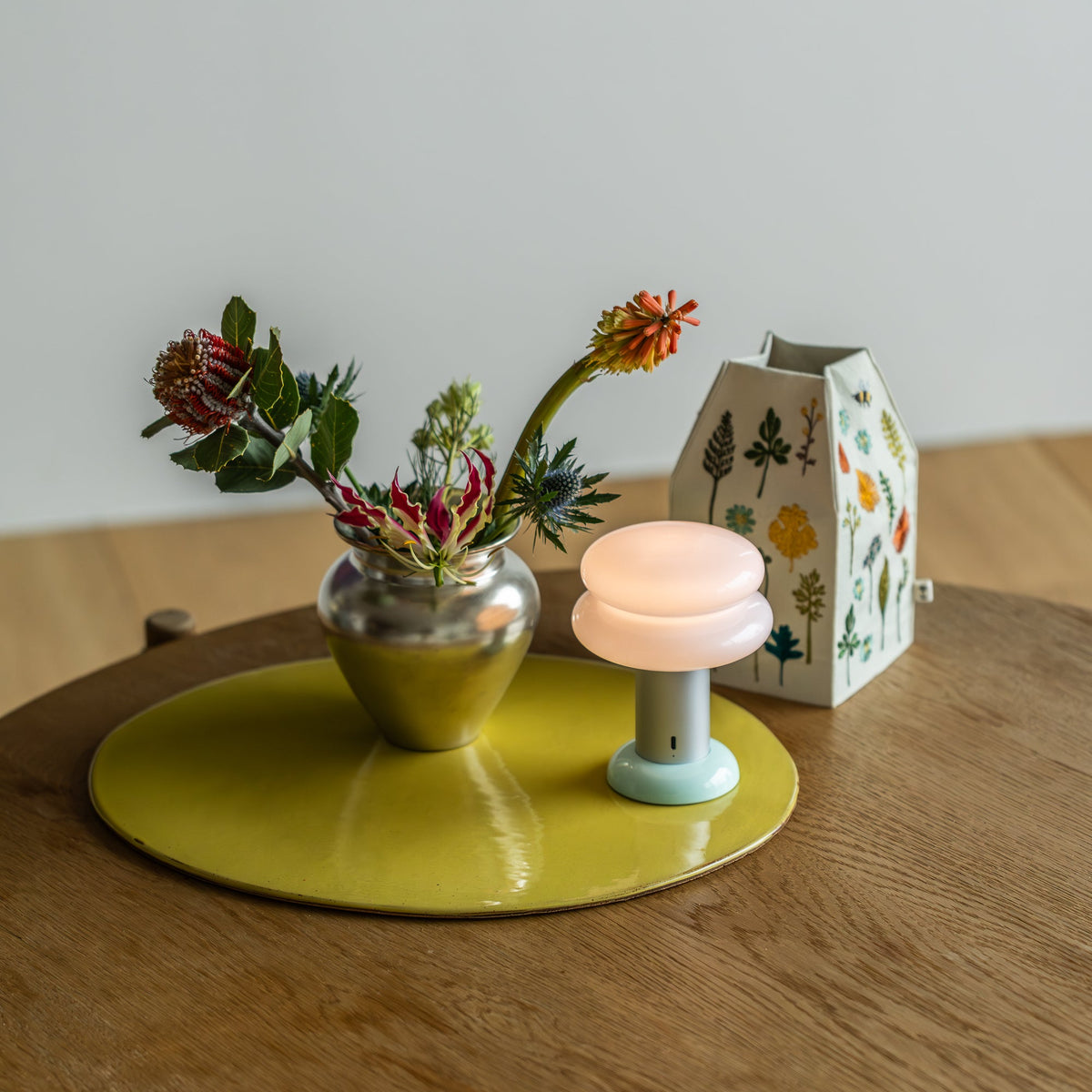 A styled interior scene showing a round wooden table with a yellow oval tray, a small glowing mushroom-shaped table lamp, a metallic vase with flowers, and a white decorative box with floral illustrations.