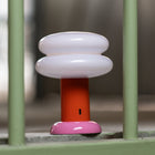 A modern mushroom-shaped table lamp with a glossy white cap, a bright orange stem, and a pink base, positioned behind a light green metal gate.
