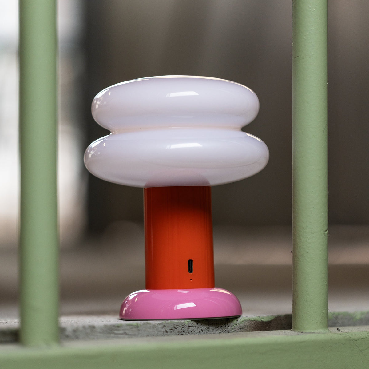 A modern mushroom-shaped table lamp with a glossy white cap, a bright orange stem, and a pink base, positioned behind a light green metal gate.