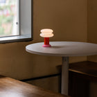 A small modern table lamp with a pink base and two stacked frosted white dome shades sits on a round white table in a room corner with a window and beige walls.