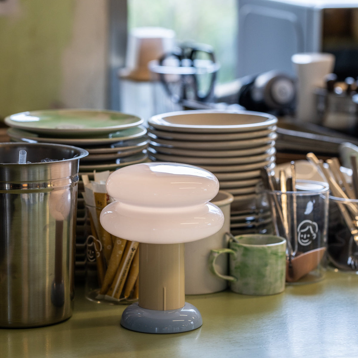 A small modern table lamp with a frosted white rounded top and a gray base sits on a glossy green countertop in a kitchen setting, surrounded by stacked plates, mugs, and metal containers.