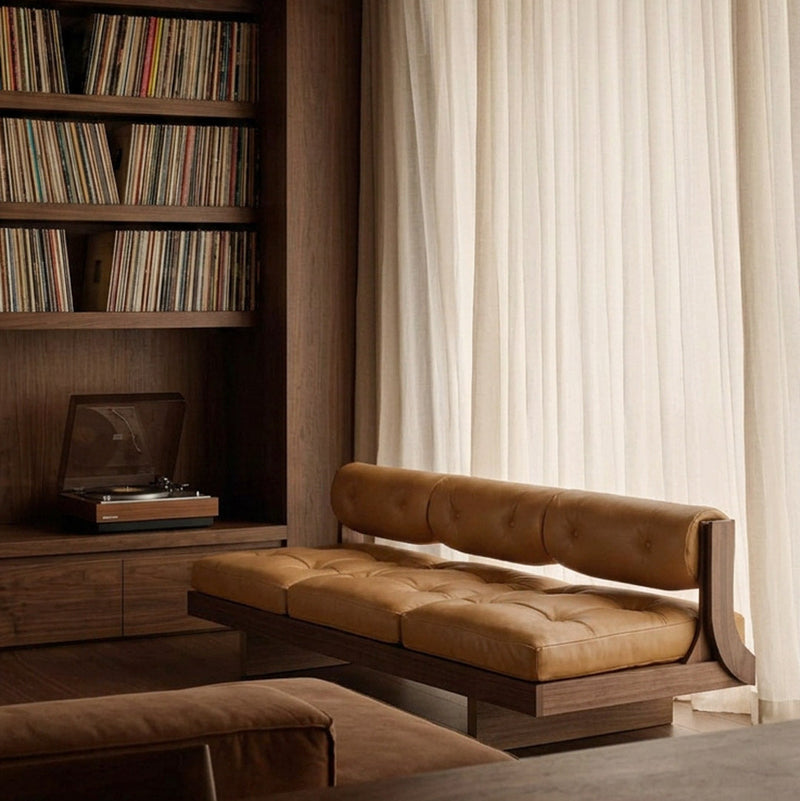 A mid-century modern tufted leather bench with a warm tan/caramel color and dark wood frame, positioned in a cozy room next to a large window with sheer white curtains and a dark wood bookshelf.
