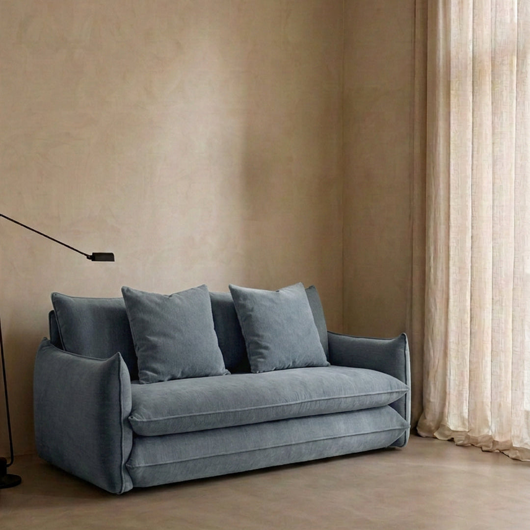 Blossom sofa bed in Celestine with curved arms and matching cushions, styled beside a floor lamp and sheer curtains.