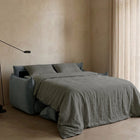 Blossom sofa bed in Celestine, unfolded and styled with grey bedding beside a curtained window.