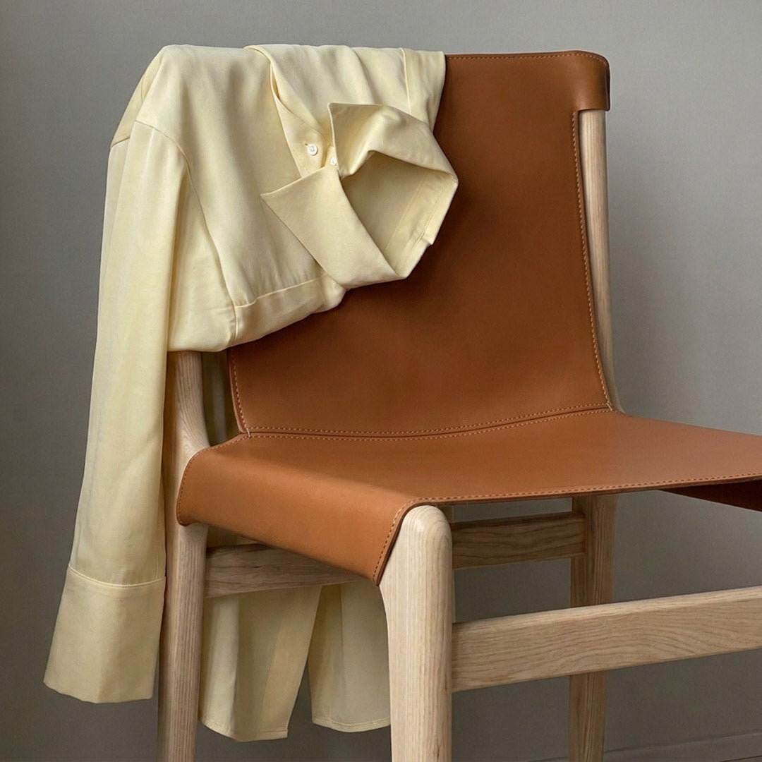 A cream-colored long-sleeve button-up shirt draped over a modern wooden chair with a tan leather seat and backrest, set against a plain gray wall.