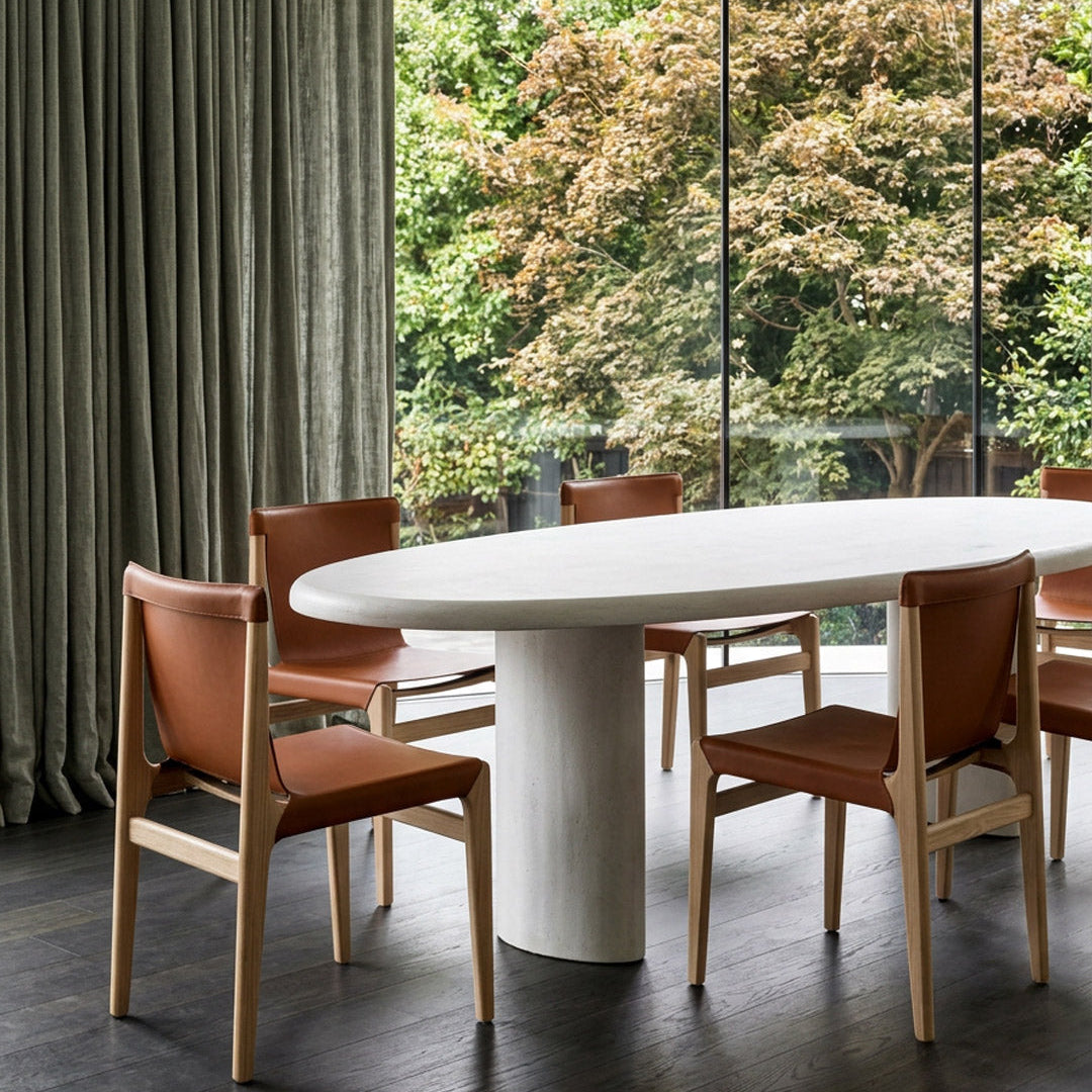 An oval white dining table with a thick cylindrical pedestal base, surrounded by four wooden dining chairs with brown leather seats, set in a modern room with a large window overlooking green trees.