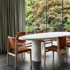 An oval white dining table with a thick cylindrical pedestal base, surrounded by four wooden dining chairs with brown leather seats, set in a modern room with a large window overlooking green trees.