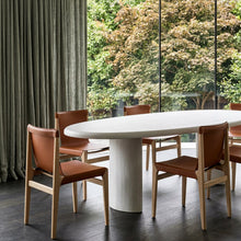 An oval white dining table with a thick cylindrical pedestal base, surrounded by four wooden dining chairs with brown leather seats, set in a modern room with a large window overlooking green trees.