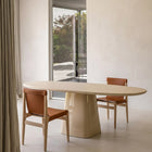 A minimalist dining room scene featuring a light beige oval table with a sculptural pedestal base, paired with two wooden chairs upholstered in warm brown leather, set against a neutral cream wall.