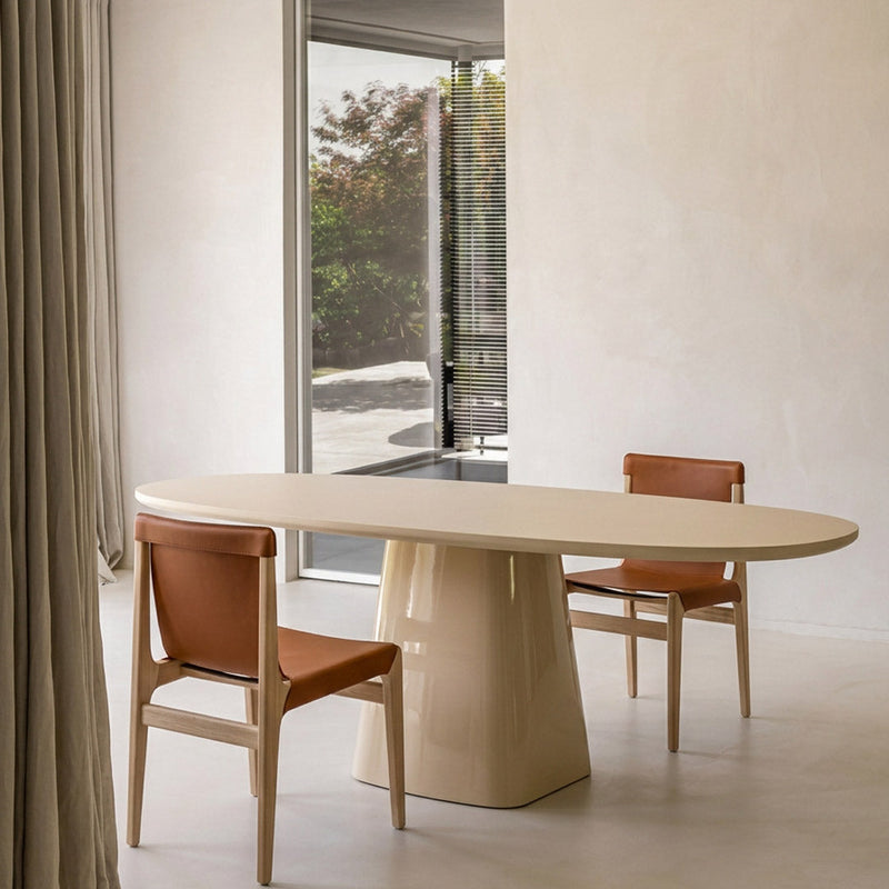 A minimalist dining room scene featuring a light beige oval table with a sculptural pedestal base, paired with two wooden chairs upholstered in warm brown leather, set against a neutral cream wall.
