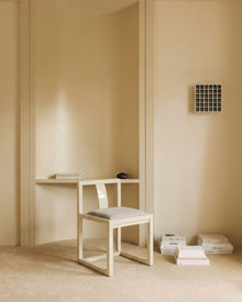 A minimalist cream-colored wooden desk and matching chair with a light gray cushion, styled in a warm, neutral-toned room with beige walls and floor.