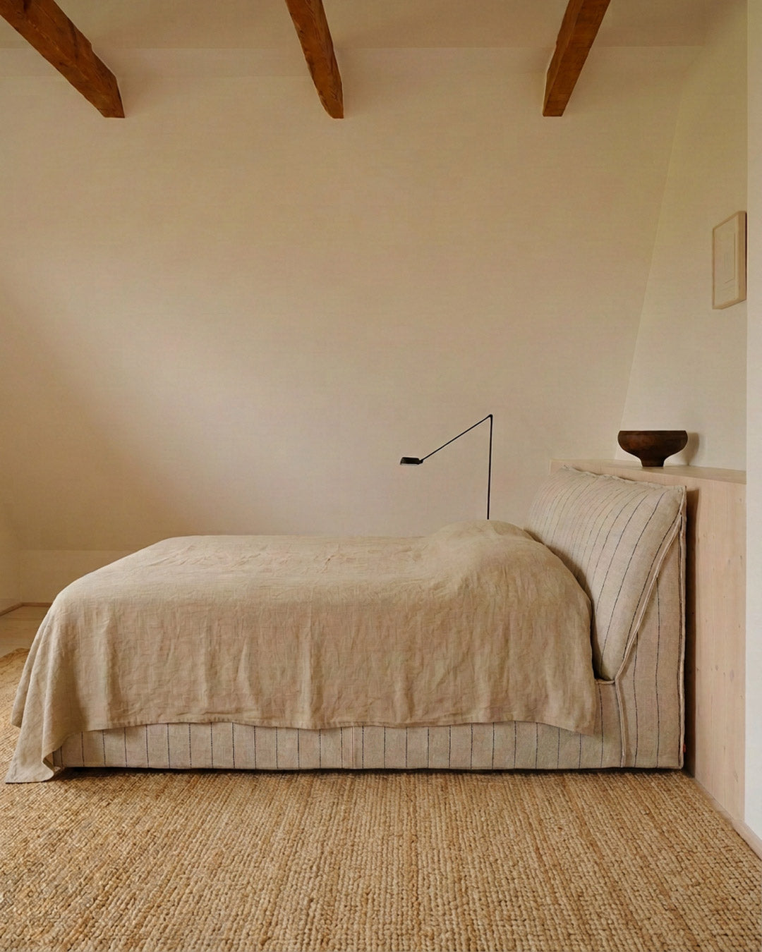 A minimalist bedroom scene featuring a bed dressed in a light beige linen cover and striped pillowcases, set against a warm cream wall with exposed wooden ceiling beams and a woven jute rug.