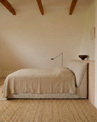 A minimalist bedroom scene featuring a bed dressed in a light beige linen cover and striped pillowcases, set against a warm cream wall with exposed wooden ceiling beams and a woven jute rug.