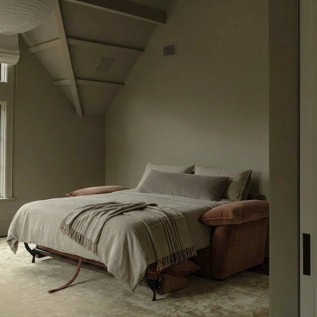 Ease sofa bed in Bronze, unfolded and styled with light bedding in a warm-toned bedroom.