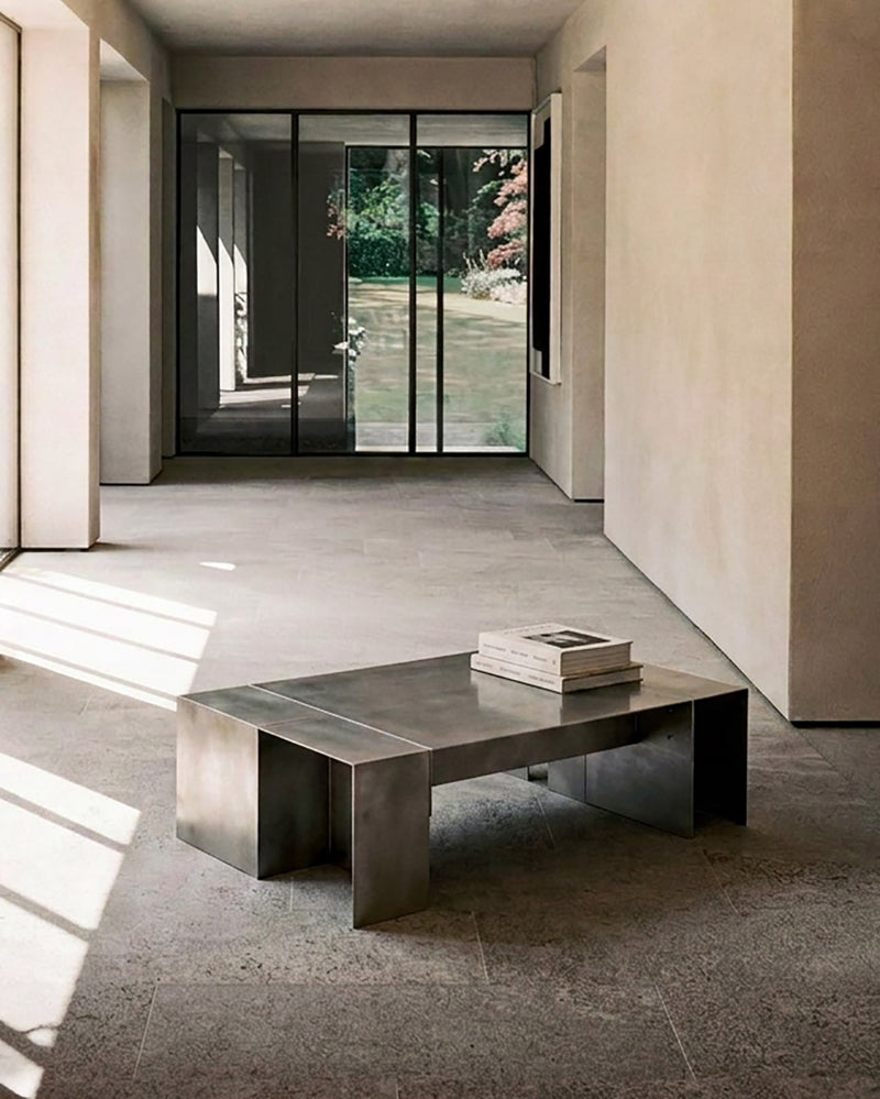 A modern rectangular metal coffee table with a brushed silver finish sits in a minimalist concrete-floored interior. A stack of books rests on top of the table, which is positioned in front of a large glass sliding door looking out onto a garden.
