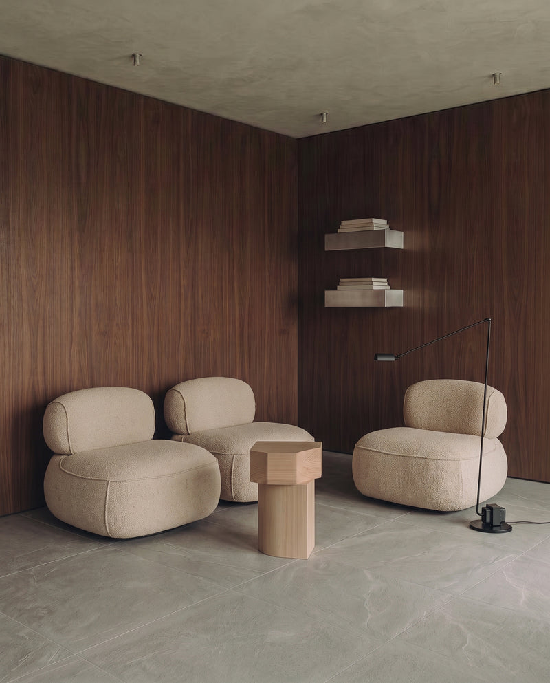 A minimalist lounge room corner featuring three beige upholstered lounge chairs, a small hexagonal wooden side table, a black floor lamp, and floating shelves against dark wood-paneled walls.