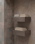 Two floating rectangular wall shelves mounted on a stone-tile wall, each holding a stack of neutral-colored folded towels or books.