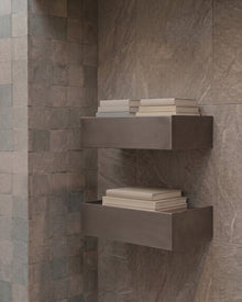 Two floating rectangular wall shelves mounted on a stone-tile wall, each holding a stack of neutral-colored folded towels or books.