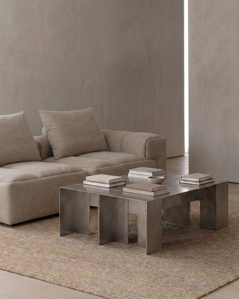 A modern rectangular coffee table with a brushed silver metallic finish, styled in a minimalist living room next to a beige sectional sofa.