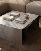 A modern square coffee table with a brushed silver metal top and dark metal legs, styled in a living room setting with books and a small bowl on top.