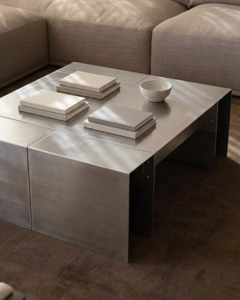 A modern square coffee table with a brushed silver metal top and dark metal legs, styled in a living room setting with books and a small bowl on top.