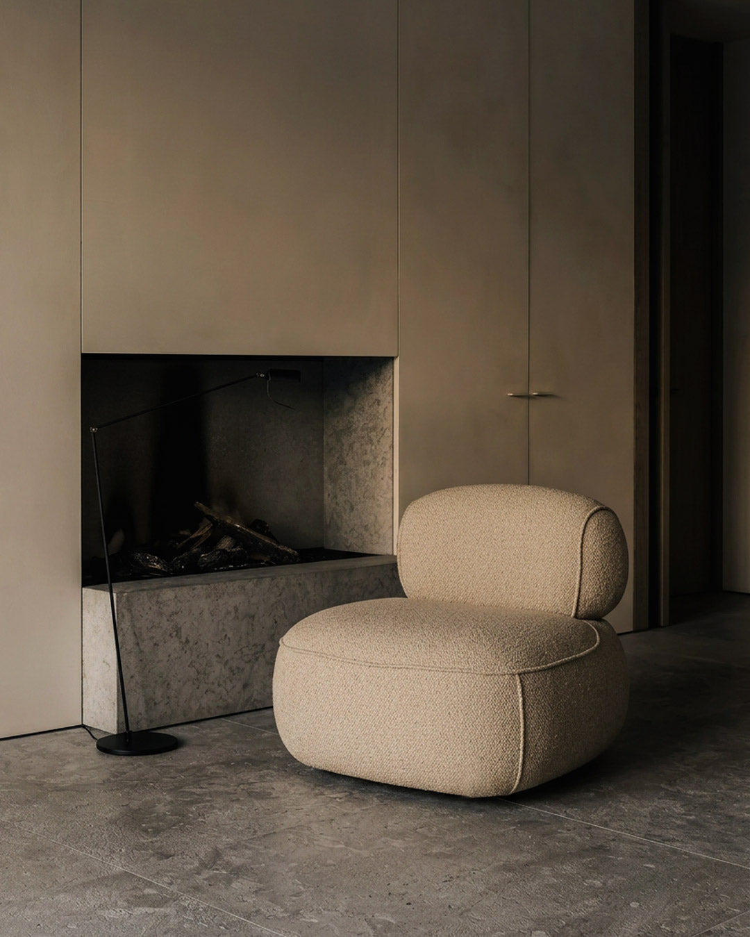 A modern low-profile armchair upholstered in textured beige bouclé fabric, positioned in a minimalist interior next to a concrete fireplace.