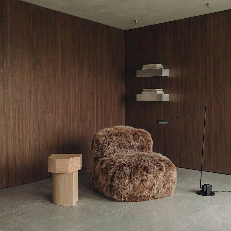 A modern room corner featuring a plush, shaggy brown armchair, a light wood side table, a black floor lamp, and dark wood-paneled walls.
