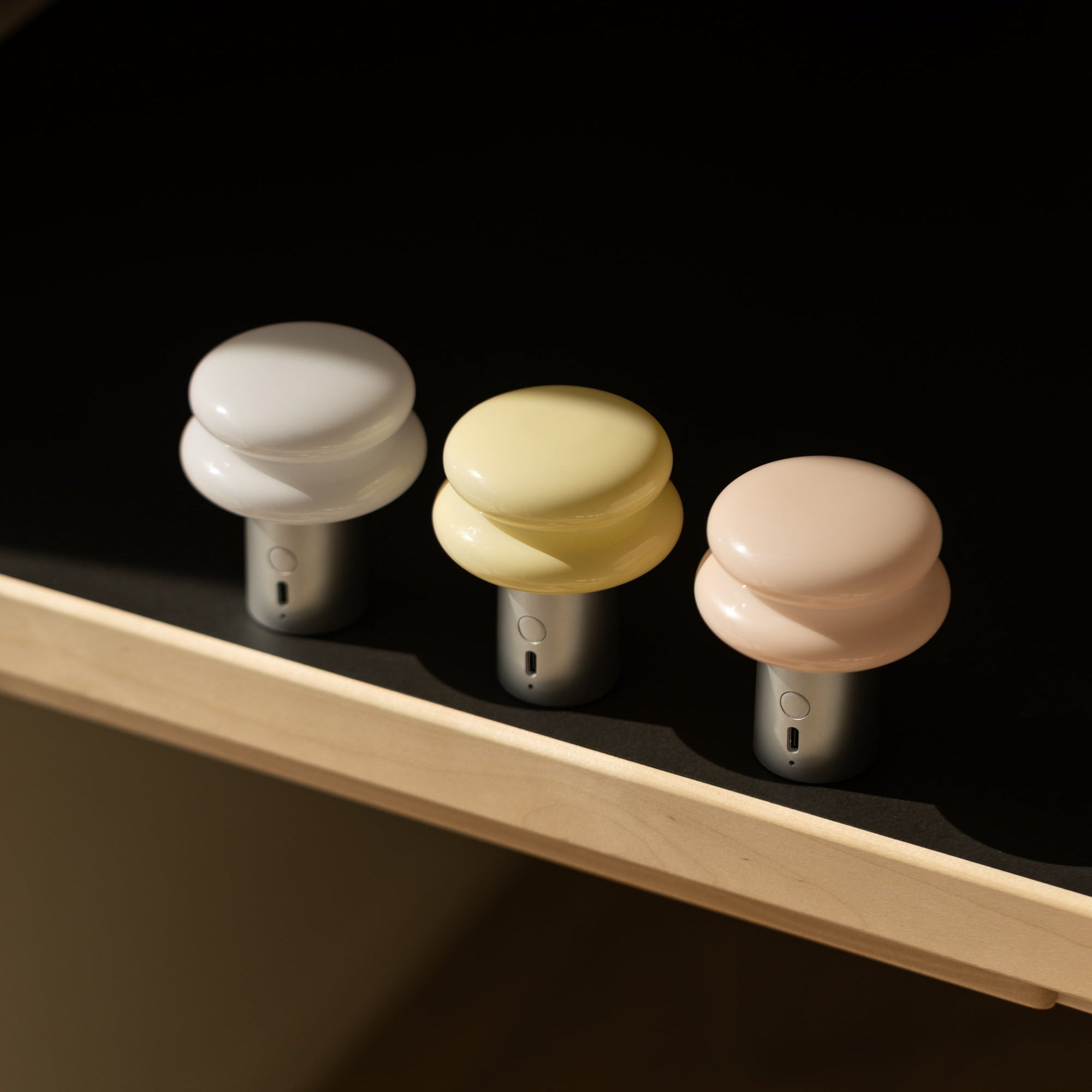 Three portable lamps with silver bases and white, butter yellow and peach/pink shades objects on a dark surface with a wooden shelf.