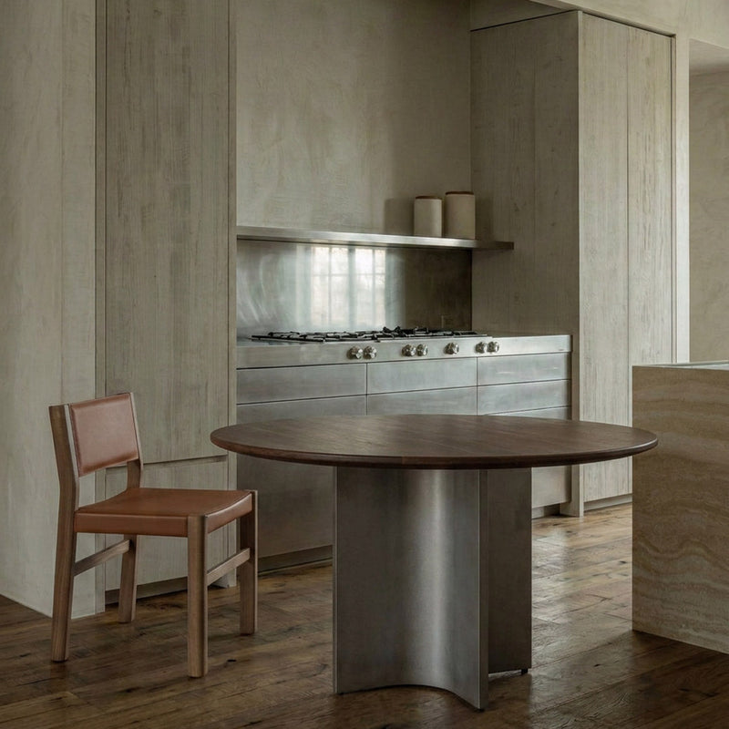 A modern kitchen interior with a round dark wood dining table in the foreground, a brown leather chair, and built-in cabinetry and a stainless steel gas range in the background.