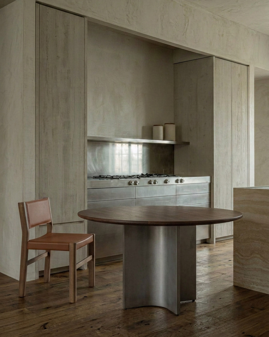 A modern kitchen interior with a round dark wood dining table in the foreground, a brown leather chair, and built-in cabinetry and a stainless steel gas range in the background.