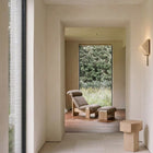 A minimalist interior hallway with warm beige walls and a large window looking out onto greenery, with a lounge chair and ottoman placed in the room beyond.
