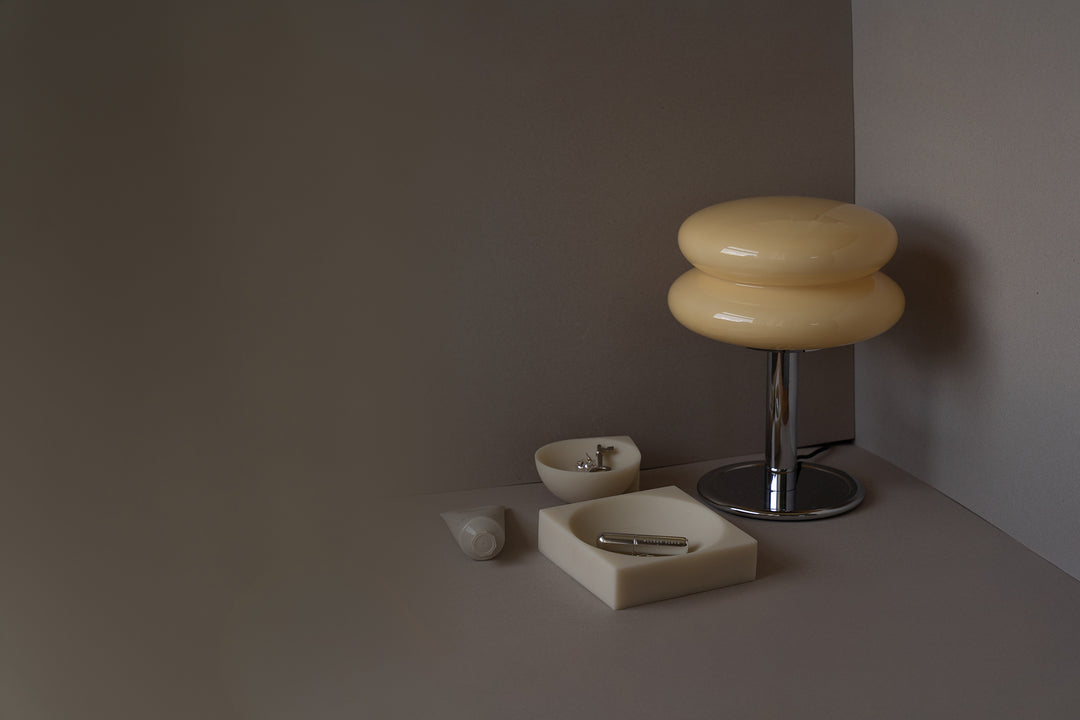 A modern table lamp with a two-tiered, rounded cream-colored shade on a chrome base, styled on a matching surface next to minimalist cream-colored ceramic accessories against a dark gray background.