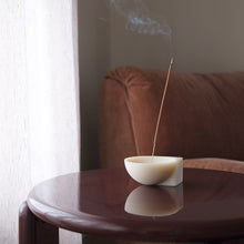 A white ceramic incense holder with a single incense stick standing inside, placed on a dark round wooden table in a softly lit room with a brown upholstered chair and curtain in the background.