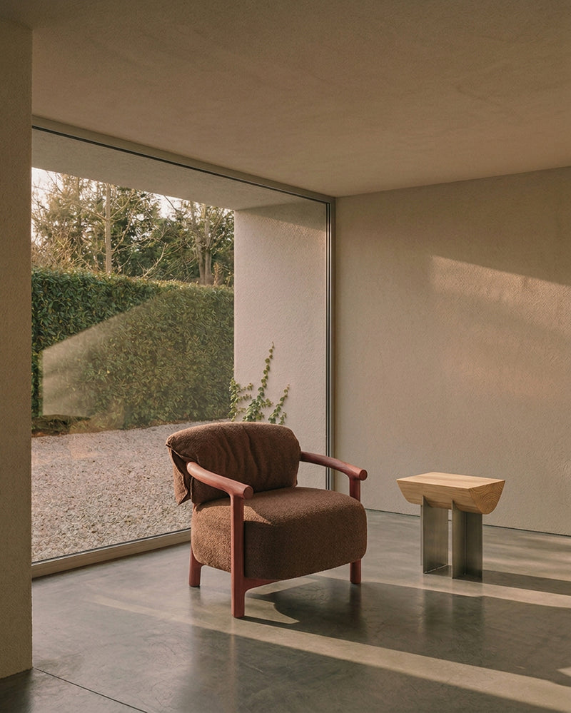 A modern brown upholstered armchair with reddish-brown wooden arms sits in a minimalist interior space with a large floor-to-ceiling window overlooking a garden. A small sculptural side table is nearby.