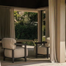 Two upholstered armchairs with dark wood frames sit on a neutral rug inside a sunlit room with large windows and curtains, overlooking a green outdoor patio.