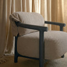 A modern armchair with a textured beige bouclé upholstered seat and back, featuring dark charcoal wood arms and legs, styled with a matching beige throw blanket draped over the arm, set against a backdrop of beige textured curtains.