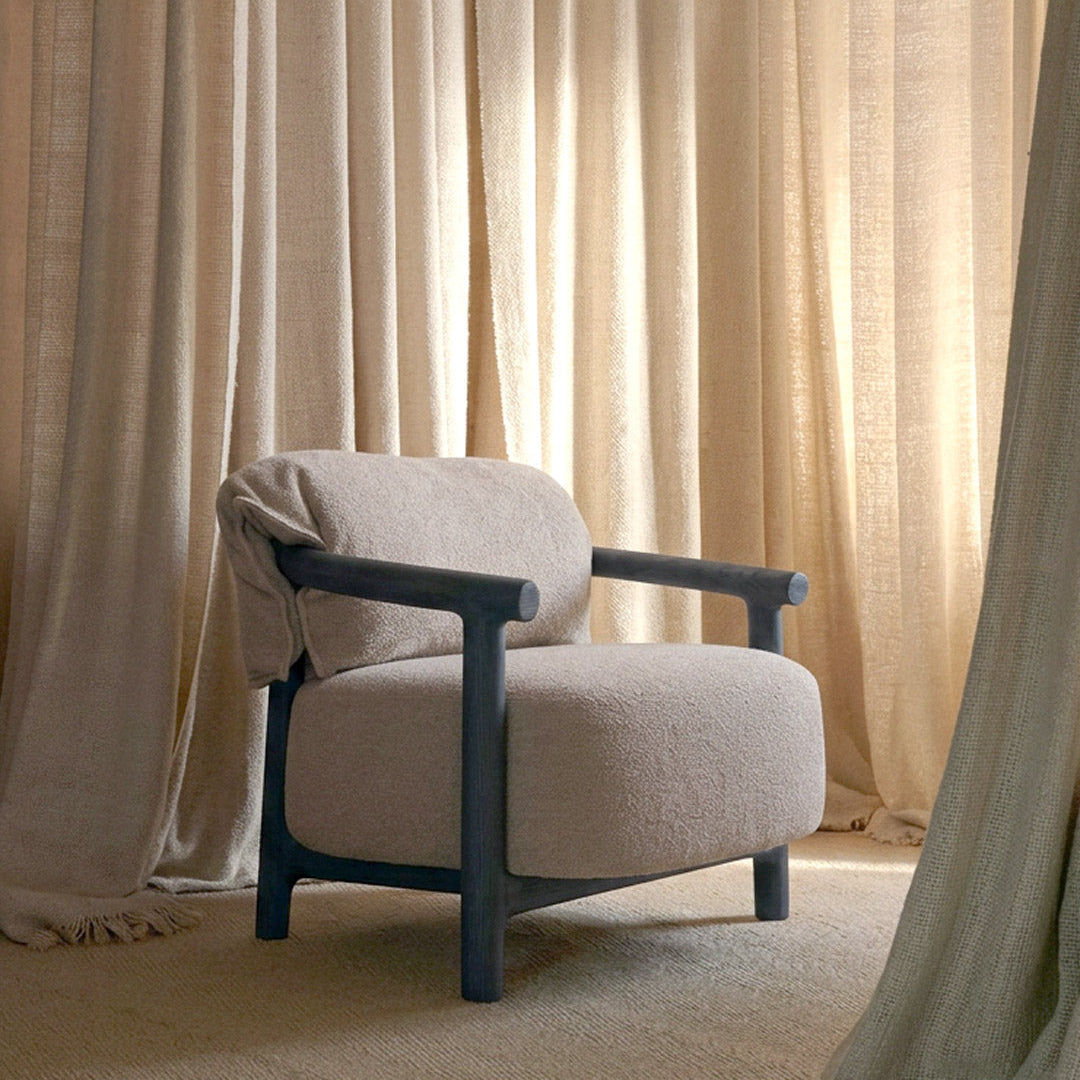 A modern upholstered armchair with a textured light beige fabric and a dark charcoal blue frame, styled in a room with beige curtains and a matching rug.