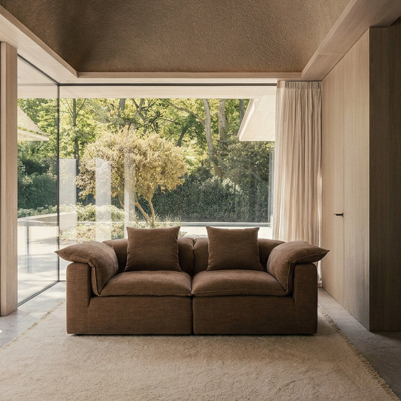 A brown upholstered two-seat sofa with two matching throw pillows, placed in a modern interior with a large window overlooking greenery, on a beige rug.