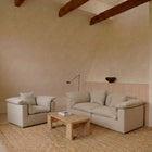 A minimalist living room setup featuring a light beige upholstered armchair and matching sofa with striped cushions, arranged on a large natural jute rug around a low wooden coffee table. The room has a warm beige plaster wall, a light wood wainscoting panel, and exposed dark wood ceiling beams.