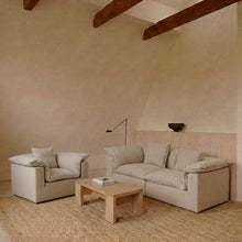 A minimalist living room setup featuring a light beige upholstered armchair and matching sofa with striped cushions, arranged on a large natural jute rug around a low wooden coffee table. The room has a warm beige plaster wall, a light wood wainscoting panel, and exposed dark wood ceiling beams.
