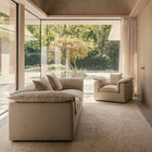 A modern beige upholstered sofa and matching armchair in a minimalist interior with large floor-to-ceiling windows overlooking a garden.