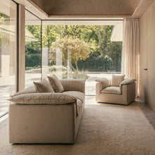 A modern beige upholstered sofa and matching armchair in a minimalist interior with large floor-to-ceiling windows overlooking a garden.