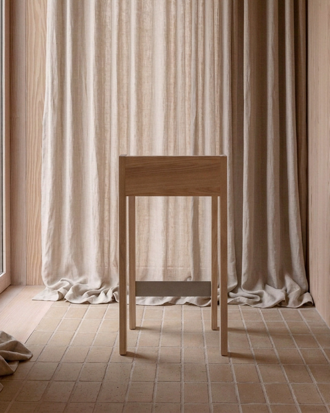 A minimalist light wood console table or stool stands in a softly lit interior with beige curtains and tiled flooring.