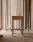 A minimalist light wood console table or stool stands in a softly lit interior with beige curtains and tiled flooring.