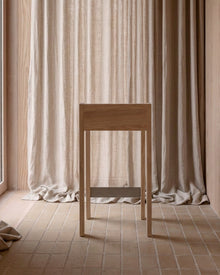 A minimalist light wood console table or stool stands in a softly lit interior with beige curtains and tiled flooring.